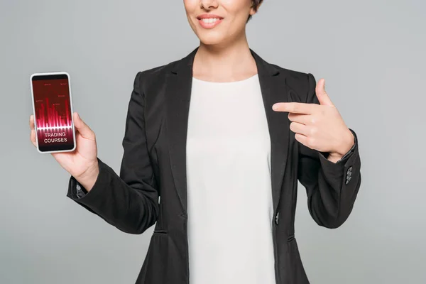 Vue partielle de souriante femme d'affaires mixte pointant du doigt le smartphone avec l'application de cours de trading à l'écran isolé sur gris — Photo de stock