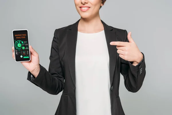 Частковий вид усміхненої змішаної раси бізнес-леді, що показує смартфон із додатком маркетингового аналізу на екрані ізольовано на сірому — стокове фото