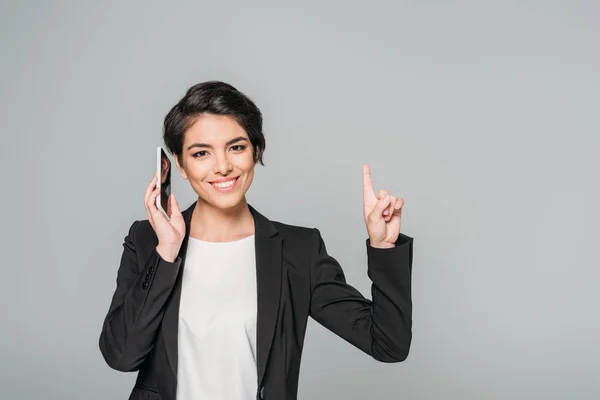 Cheerful mixed race businesswoman talking on smartphone and showing idea gesture isolated on grey — Stock Photo