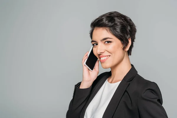 Feliz mixta mujer de negocios hablando en el teléfono inteligente y sonriendo a la cámara aislada en gris - foto de stock