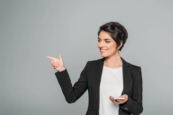 Sonriente mujer de negocios de raza mixta sosteniendo teléfono inteligente y apuntando con el dedo aislado en gris - foto de stock
