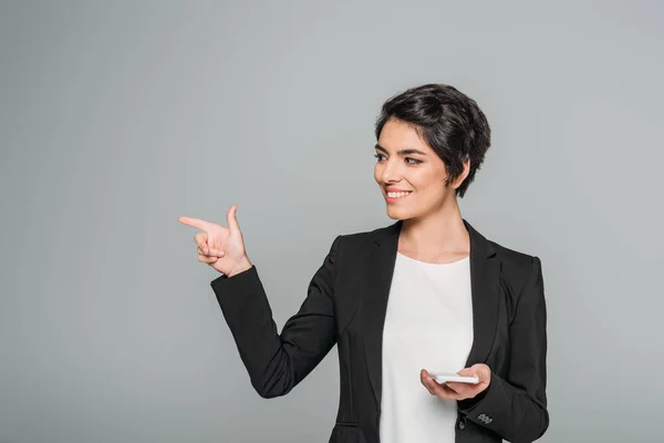 Sorridente donna d'affari mista che tiene lo smartphone e punta con il dito isolato sul grigio — Foto stock