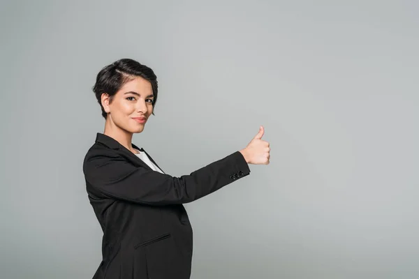 Усміхнена змішана гонка бізнес-леді показує великий палець і дивиться на камеру ізольовано на сірому — стокове фото