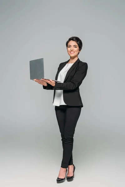 Atractiva mujer de negocios de raza mixta sosteniendo portátil y sonriendo a la cámara sobre fondo gris - foto de stock