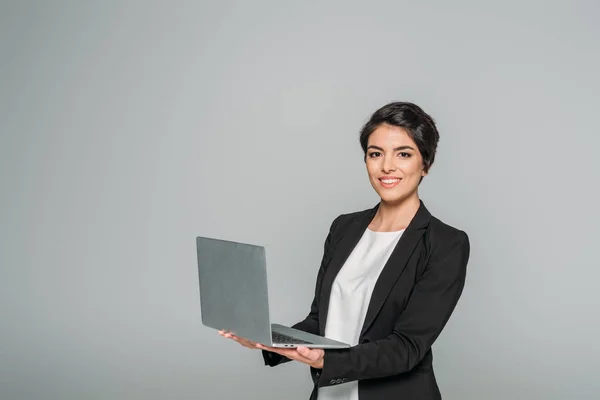 Весела змішана гонка бізнес-леді тримає ноутбук, посміхаючись на камеру ізольовано на сірому — стокове фото
