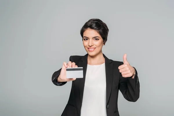 Sonriente mestiza empresaria sosteniendo tarjeta de crédito y mostrando el pulgar hacia arriba aislado en gris - foto de stock