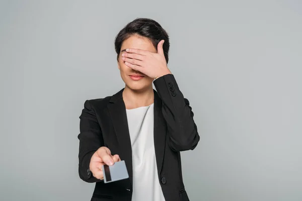 Joven mujer de negocios de raza mixta cubriendo la cara con la mano mientras sostiene la tarjeta de crédito en la mano extendida aislado en gris - foto de stock