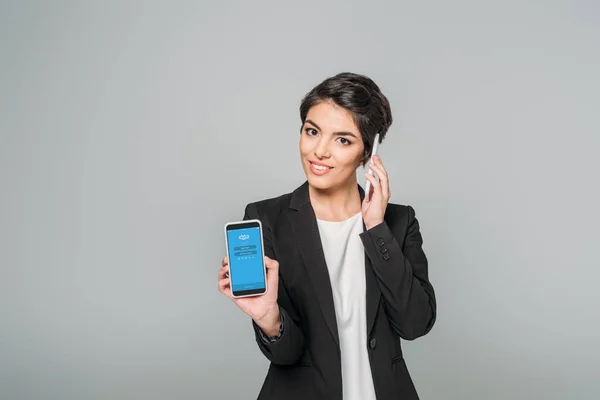 KYIV, UCRANIA - 24 DE ABRIL DE 2019: Atractiva mujer de negocios de raza mixta mostrando el teléfono inteligente con la aplicación de Skype en la pantalla y hablando en un teléfono inteligente aislado en gris . - foto de stock