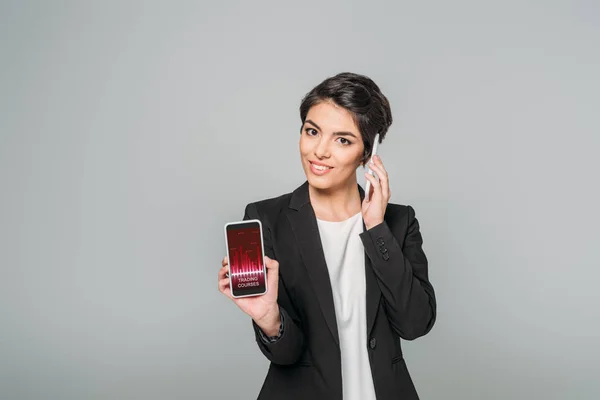 Hermosa mujer de negocios de raza mixta que muestra el teléfono inteligente con la aplicación de cursos de comercio en pantalla mientras habla en el teléfono inteligente aislado en gris - foto de stock