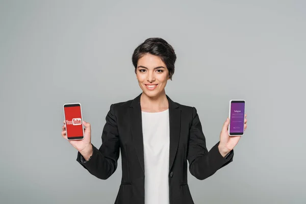 KYIV, UCRANIA - 24 DE ABRIL DE 2019: La alegre empresaria de raza mixta sostiene teléfonos inteligentes con aplicaciones de Youtube e Instagram en pantalla aislada en gris . - foto de stock