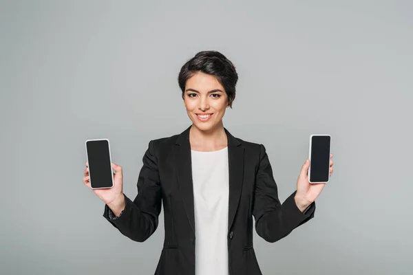 Alegre mujer de negocios de raza mixta mostrando teléfonos inteligentes con pantalla en blanco aislado en gris - foto de stock
