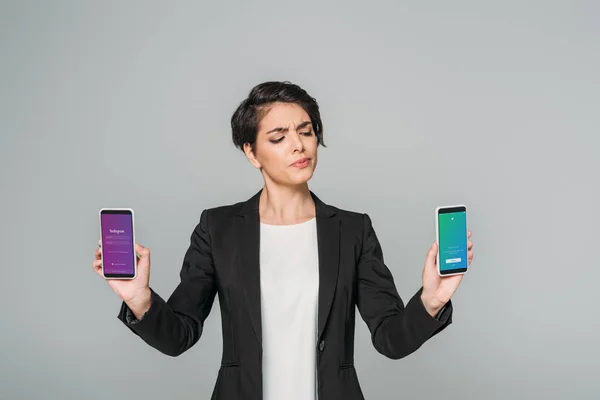 KYIV, UCRANIA - 24 DE ABRIL DE 2019: Empresaria escéptica de raza mixta que sostiene teléfonos inteligentes con aplicaciones de Instagram y Twitter en pantalla aislada en gris . - foto de stock