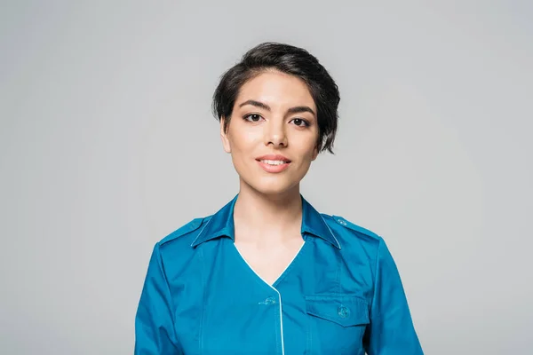 Pretty mixed race nurse in uniform smiling at camera isolated on grey — Stock Photo