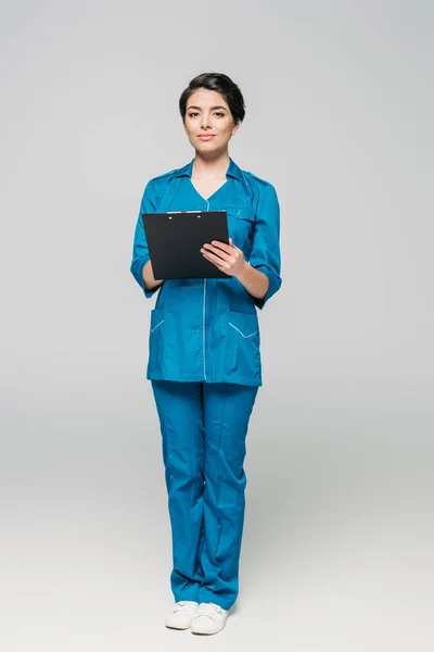 Young mixed race doctor holding clipboard while looking at camera on grey background — Stock Photo