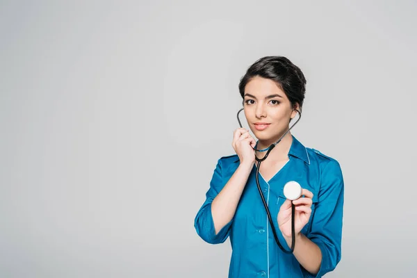 Pretty mixed race nurse using stethoscope while looking at camera isolated on grey — Stock Photo