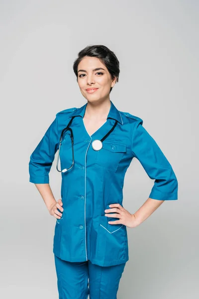Attractive mixed race doctor posing at camera with hands on hips isolated on grey — Stock Photo