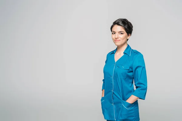 Beautiful mixed race nurse holding hands in pockets and looking at camera isolated on grey — Stock Photo