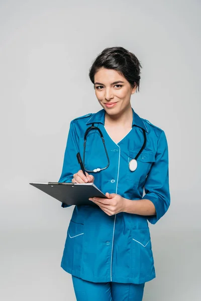 Allegro medico di razza mista con scrittura stetoscopio sugli appunti mentre guarda la fotocamera isolata sul grigio — Foto stock