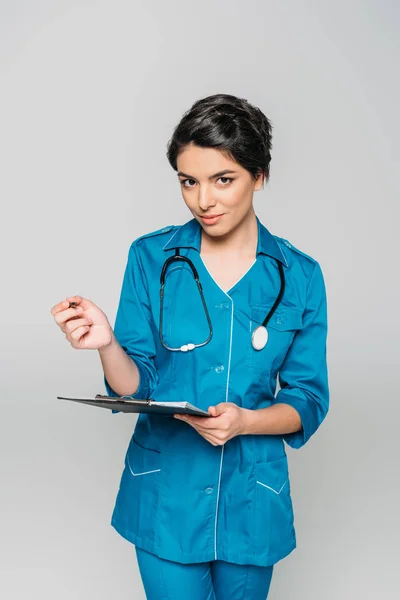 Attractive mixed race doctor writing on clipboard and looking at camera isolated on grey — Stock Photo