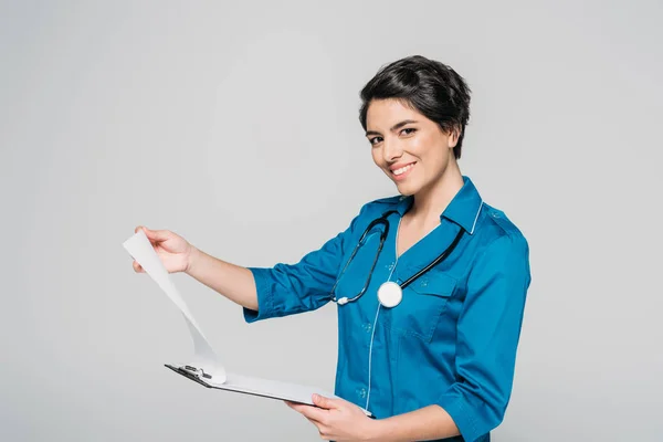 Cheerful mixed race doctor holding clipboard with papers and looking at camera isolated on grey — Stock Photo