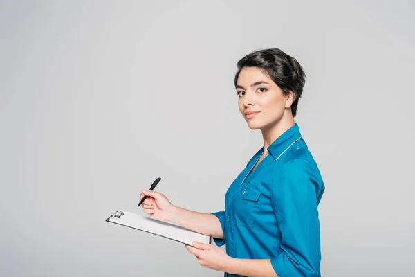 Attractive nurse in uniform writing ob clipboard and looking at camera isolated on grey — Stock Photo