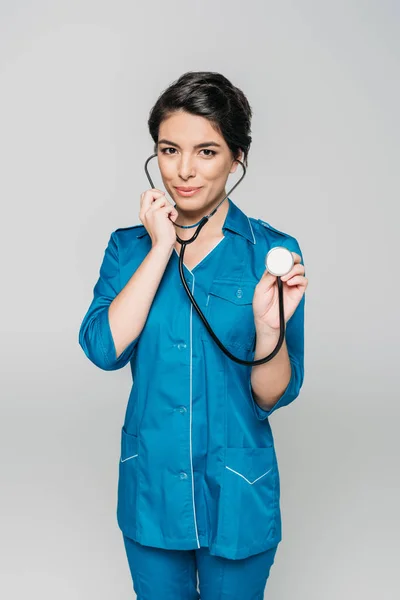 Smiling mixed race nurse using stethoscope while looking at camera isolated on grey — Stock Photo