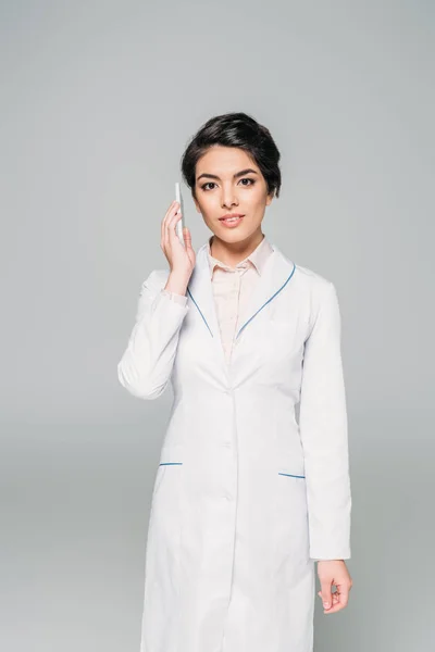Pretty mixed race doctor talking on smartphone and looking at camera isolated on grey — Stock Photo