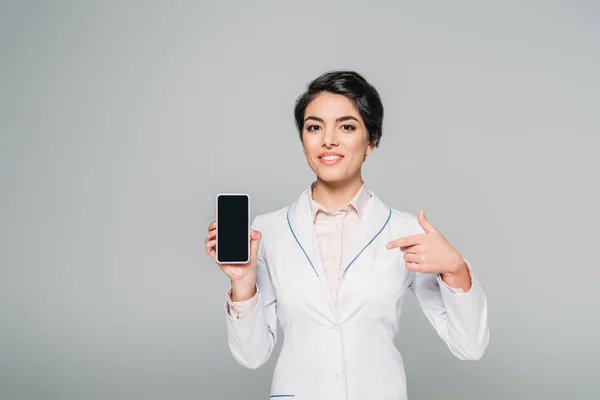 Alegre médico de raza mixta apuntando con el dedo al teléfono inteligente con pantalla en blanco aislado en gris - foto de stock