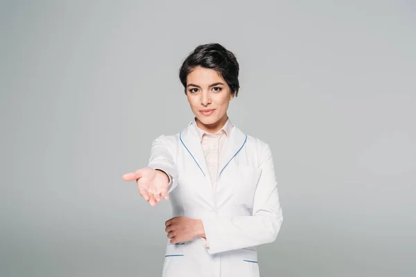 Attraente medico di razza mista guardando la fotocamera con mano sollevata isolato su grigio — Foto stock