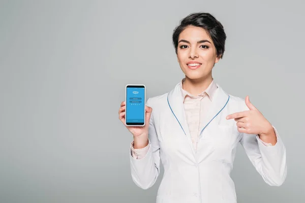KYIV, UCRANIA - 24 DE ABRIL DE 2019: Bastante médico de raza mixta señalando con el dedo al teléfono inteligente con la aplicación de Skype en la pantalla aislada en gris . - foto de stock