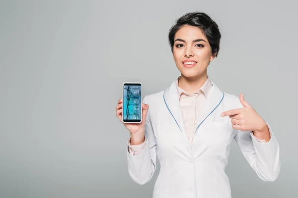 Sonriente médico de raza mixta apuntando con el dedo al teléfono inteligente con prueba de ADN en la pantalla aislado en gris - foto de stock