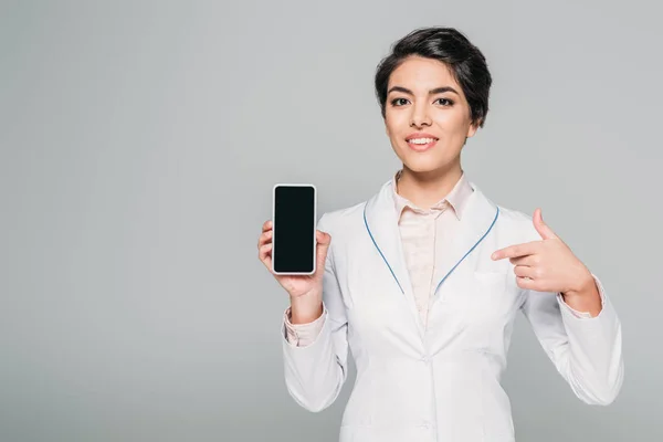 Alegre médico de raza mixta mostrando teléfono inteligente con pantalla en blanco aislado en gris - foto de stock