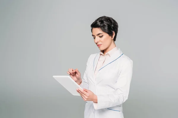 Attraktiver Mischlingsarzt im weißen Kittel mit digitalem Tablet isoliert auf grau — Stockfoto