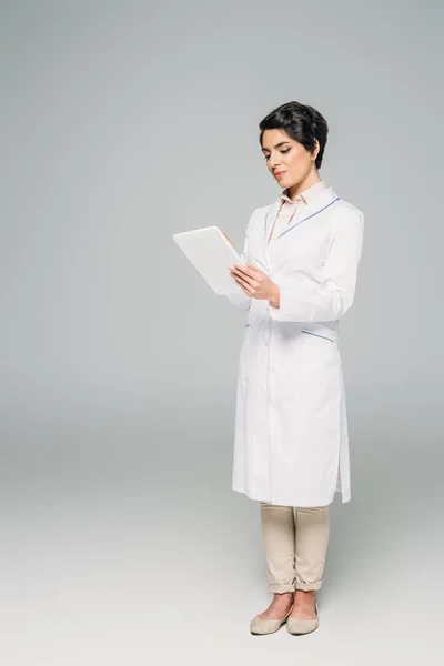 Красивый врач смешанной расы в белом халате с помощью цифрового планшета на сером фоне — стоковое фото