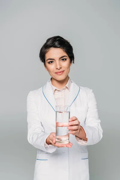 Atractivo médico de raza mixta sosteniendo un vaso de agua y mirando a la cámara aislada en gris - foto de stock