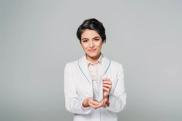 Pretty mixed race doctor holding glass of water while looking at camera isolated on grey — Stock Photo