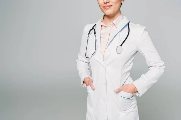 Partial view of mixed race doctor with stethoscope posing at camera with hands in pockets isolated on grey — Stock Photo