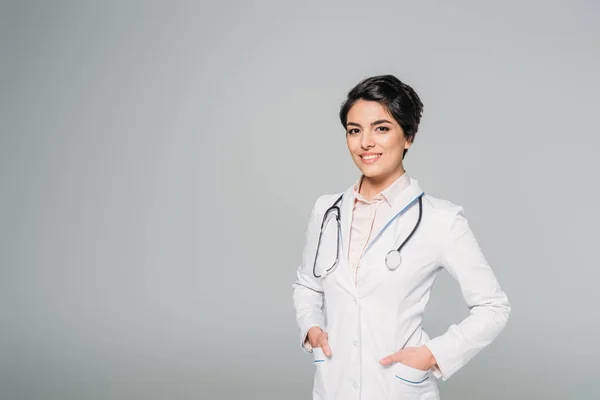 Cheerful mixed race doctor with stethoscope posing at camera with hands in pockets isolated on grey — Stock Photo