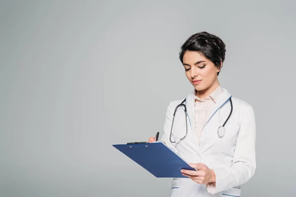 Concentrated mixed race doctor writing on clipboard isolated on grey — Stock Photo