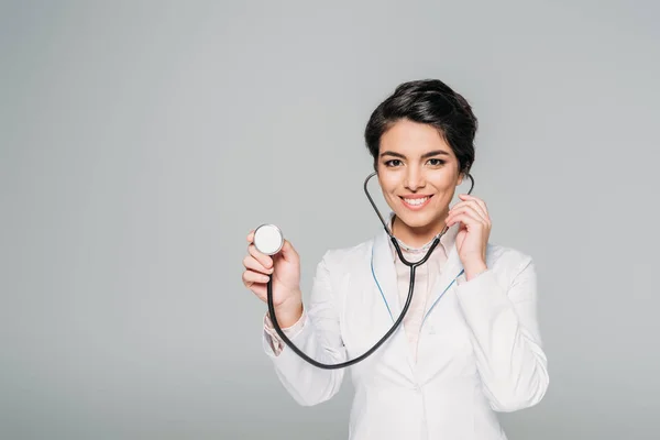 Cheerful mixed race doctor in white coat using stethoscope isolated on grey — Stock Photo