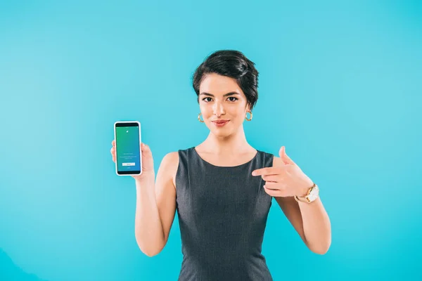 KYIV, UCRANIA - 24 DE ABRIL DE 2019: Bastante mujer de raza mixta señalando con el dedo al teléfono inteligente con la aplicación de Twitter en la pantalla aislada en azul . - foto de stock