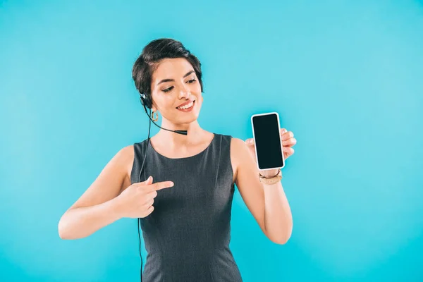 Sonriente operador de centro de llamadas de raza mixta apuntando con el dedo en el teléfono inteligente con pantalla en blanco aislado en azul - foto de stock