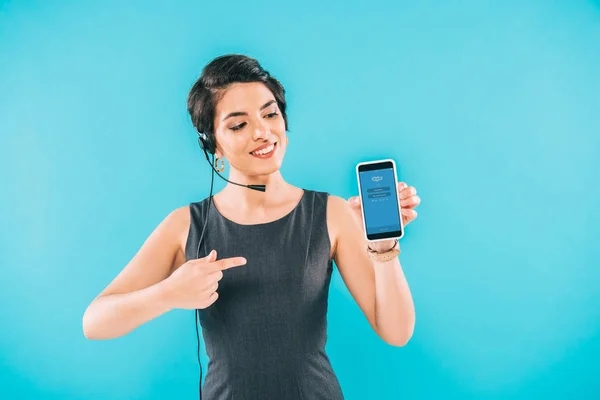 KYIV, UKRAINE - APRIL 24, 2019: Cheerful mixed race call center operator presenting smartphone with Skype app on screen isolated on blue. — Stock Photo