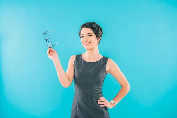 Alegre mixta carrera mujer de negocios sosteniendo gafas mientras posando en la cámara con la mano en la cadera aislado en azul — Stock Photo