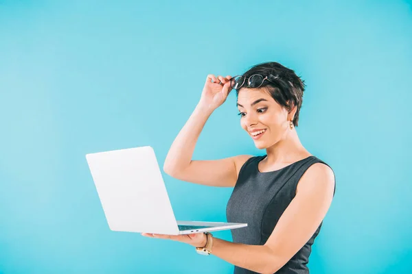 Sorprendida mujer de negocios de raza mixta tocar gafas mientras se utiliza el ordenador portátil aislado en azul - foto de stock