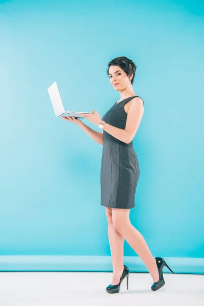 Moda mixta carrera mujer de negocios celebración de la computadora portátil y mirando a la cámara sobre fondo azul - foto de stock