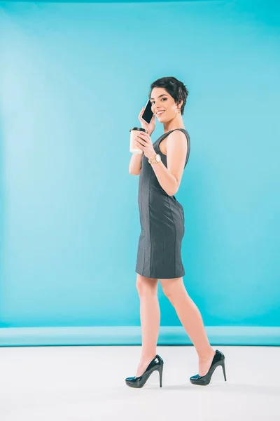 Mujer de raza mixta de moda posando en la cámara mientras habla en el teléfono inteligente y sosteniendo la taza de papel sobre fondo azul - foto de stock