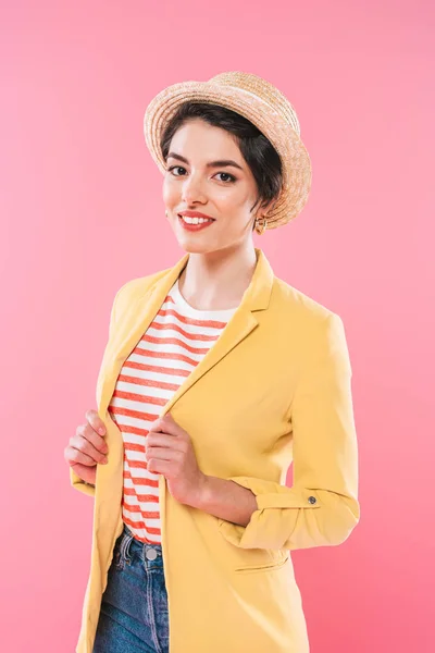 Atractiva mujer de raza mixta en ropa brillante y sombrero de paja sonriendo a la cámara aislada en rosa - foto de stock