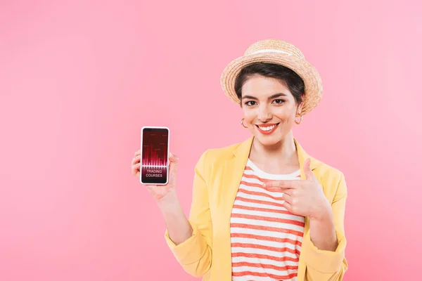 Alegre mujer de raza mixta señalando con el dedo en el teléfono inteligente con cursos de comercio aplicación en la pantalla aislada en rosa - foto de stock