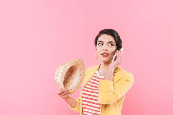 Bastante mujer de raza mixta hablando en el teléfono inteligente y sosteniendo sombrero de paja aislado en rosa - foto de stock